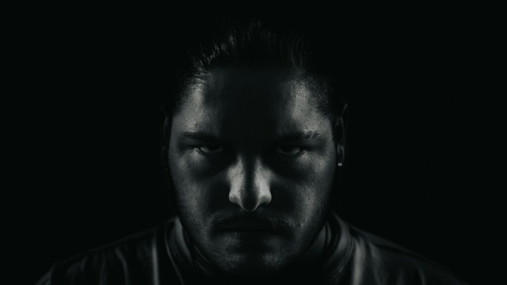 Close-up of a man's face in low lighting, with shadows obscuring most features and a focused, intense gaze from the eyes.