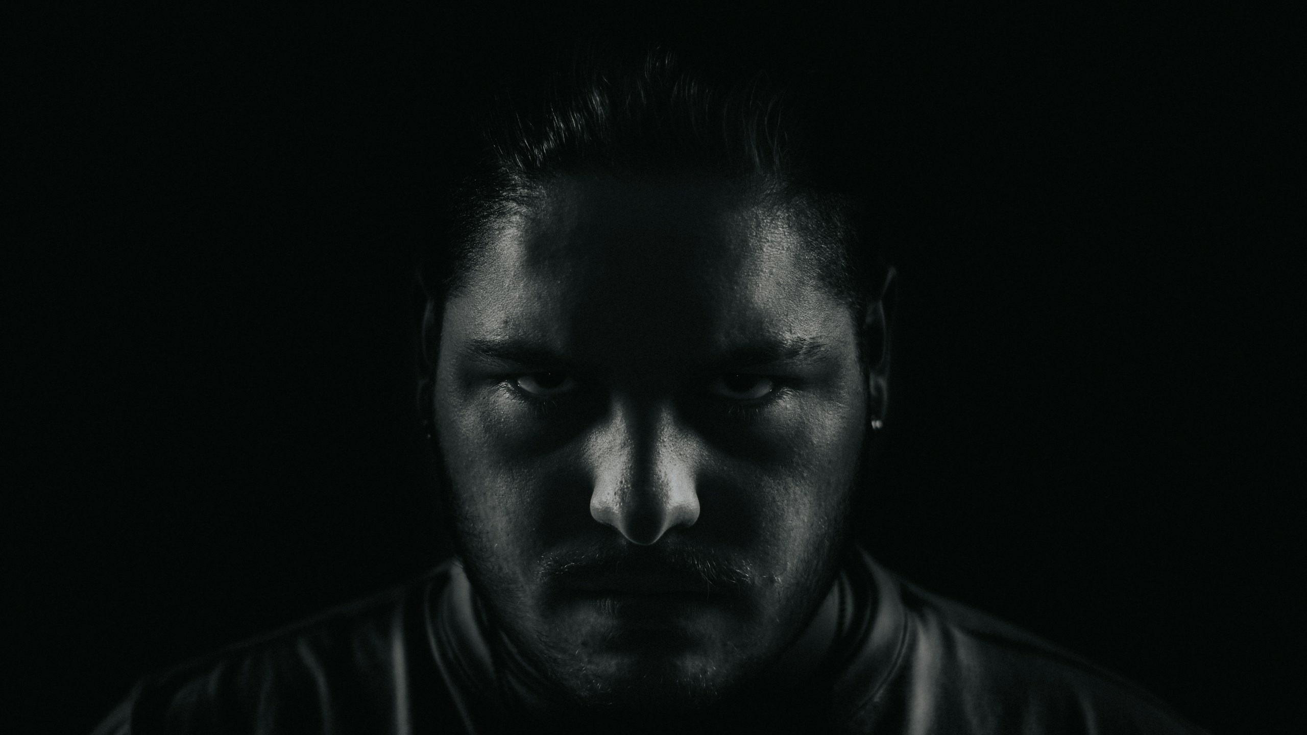 Close-up of a man's face in low lighting, with shadows obscuring most features and a focused, intense gaze from the eyes.