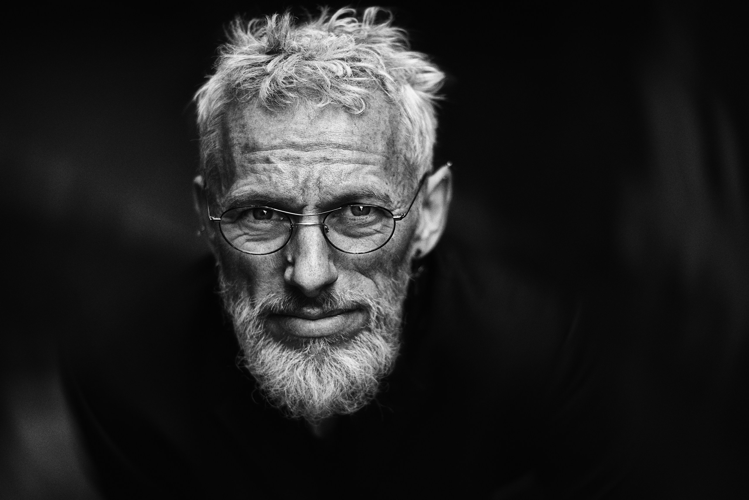 Close-up black-and-white portrait of an older man with gray hair, beard, and round glasses looking at the camera.